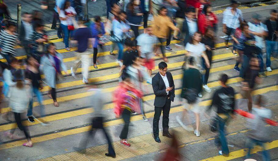 A man standing in the middle of people walking on a busy sidewalk while looking at his phone.